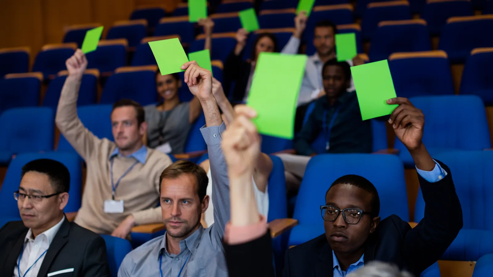 Audience members at a business meeting raise green voting cards.