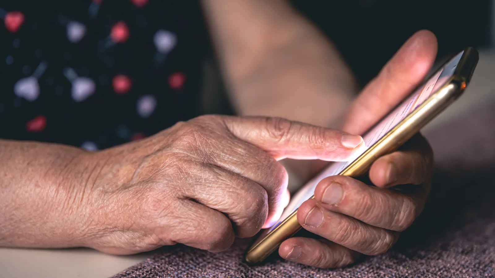 Close-up of an older person's hands tapping the screen of a gold smartphone resting on a knit blanket.