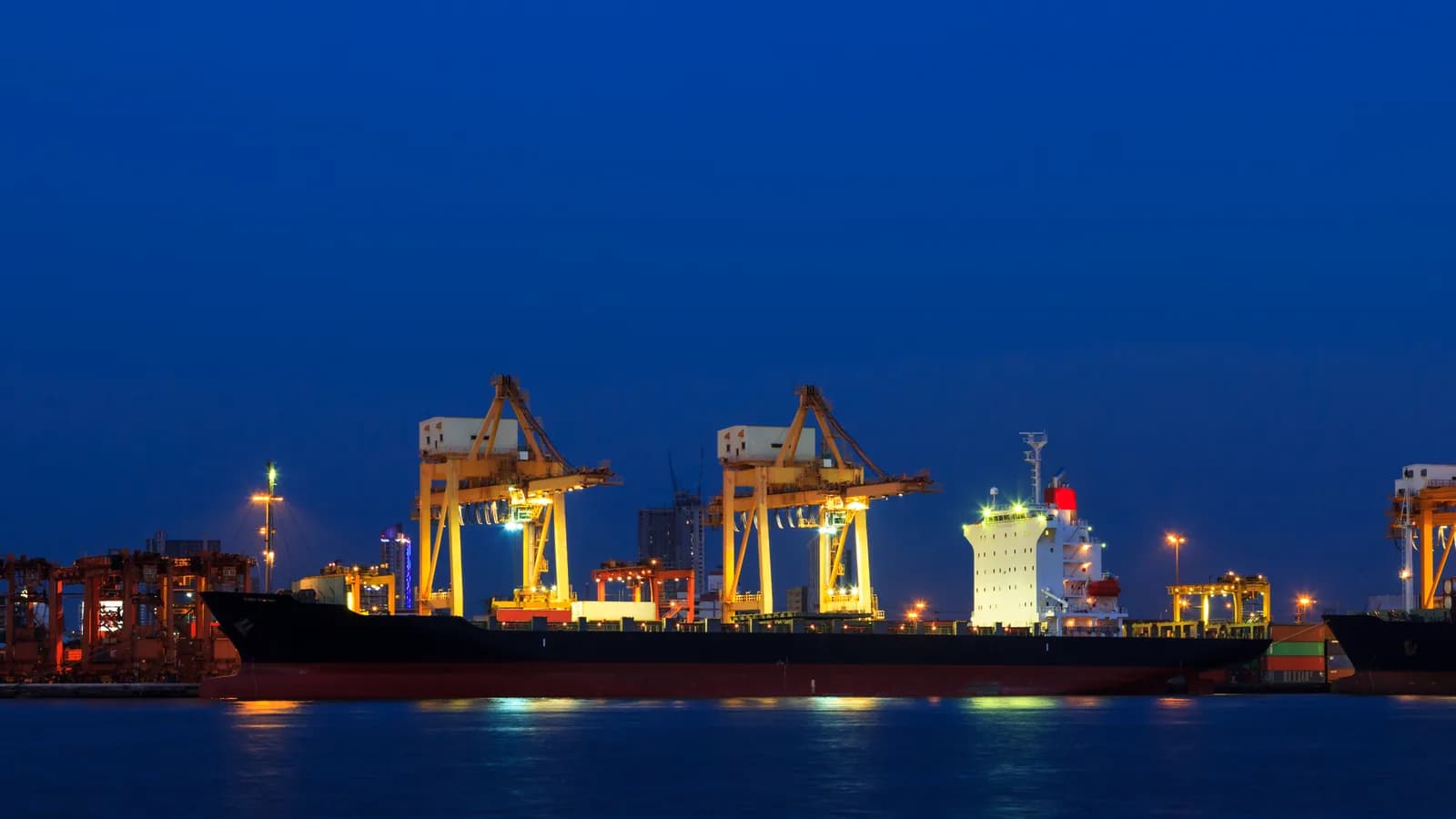 A loaded container ship docked beneath lit gantry cranes at a cargo port at twilight, with the lights reflected in dark blue water.
