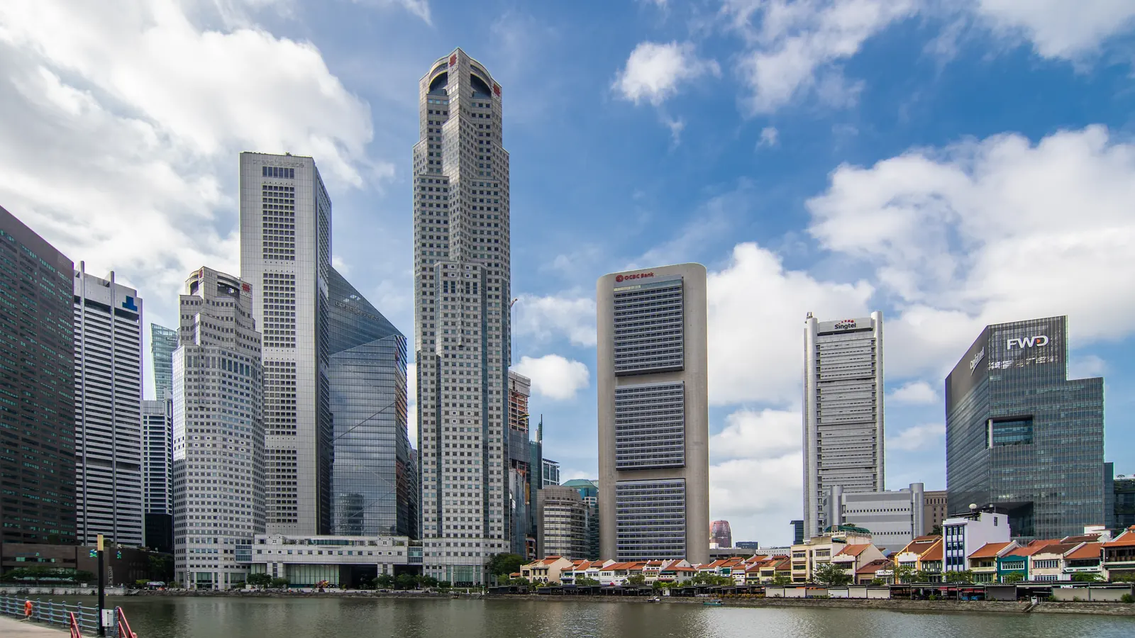Singapore's downtown financial district towers rise above the Singapore River under a blue sky with white clouds.